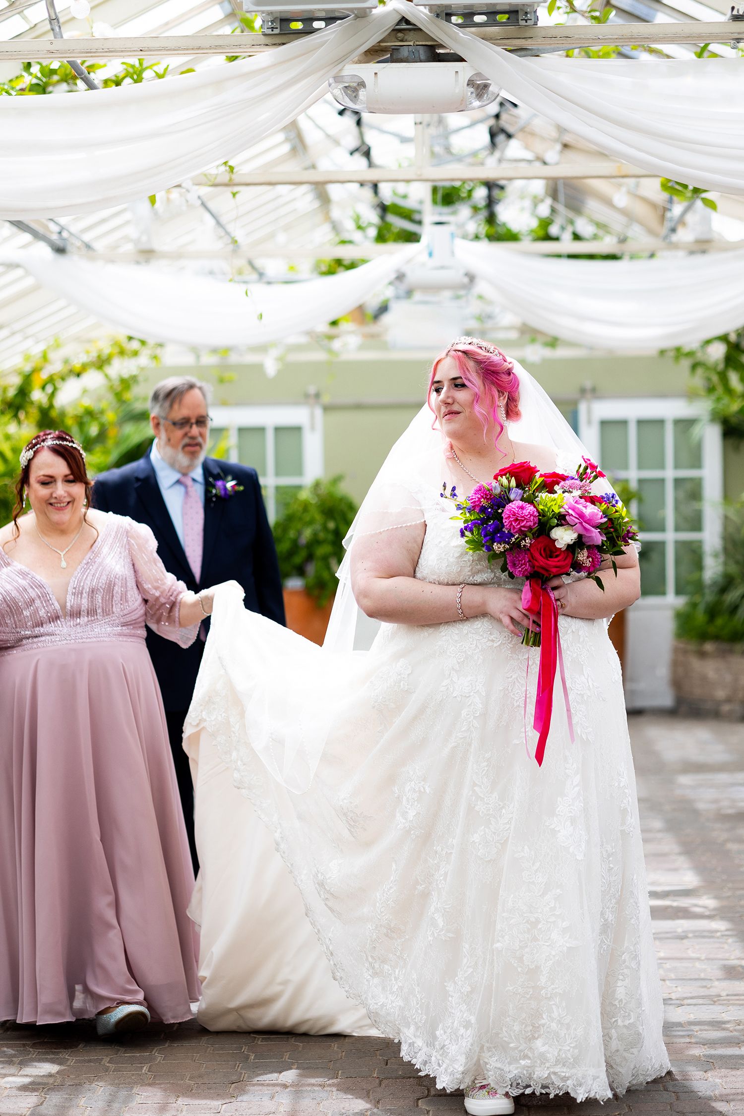 Bride with pink hair holding colorful bouquet walks down aisle in white lace gown, assisted by bridesmaid in lavender dress.