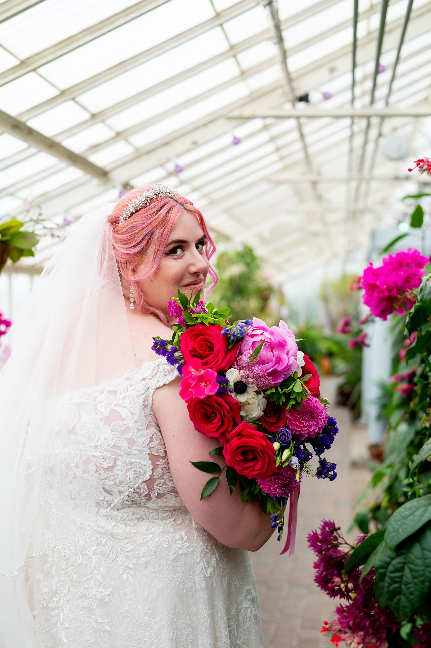 Bride with pink hair holding vibrant bouquet of red, pink, and purple flowers in a greenhouse.