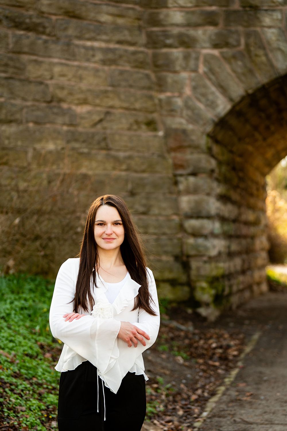A person in a white blouse and dark pants stands smiling in front of a stone archway tunnel.