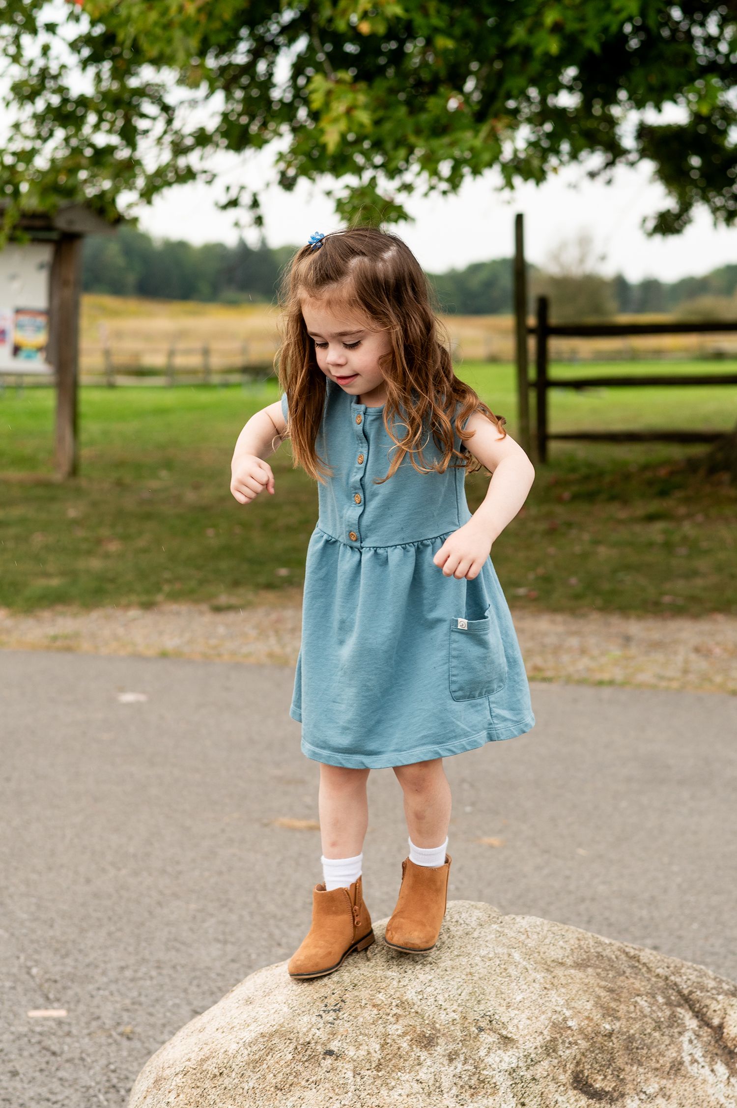 A young girl in a teal dress and brown boots stands on a rock outdoors near a wooden fence.