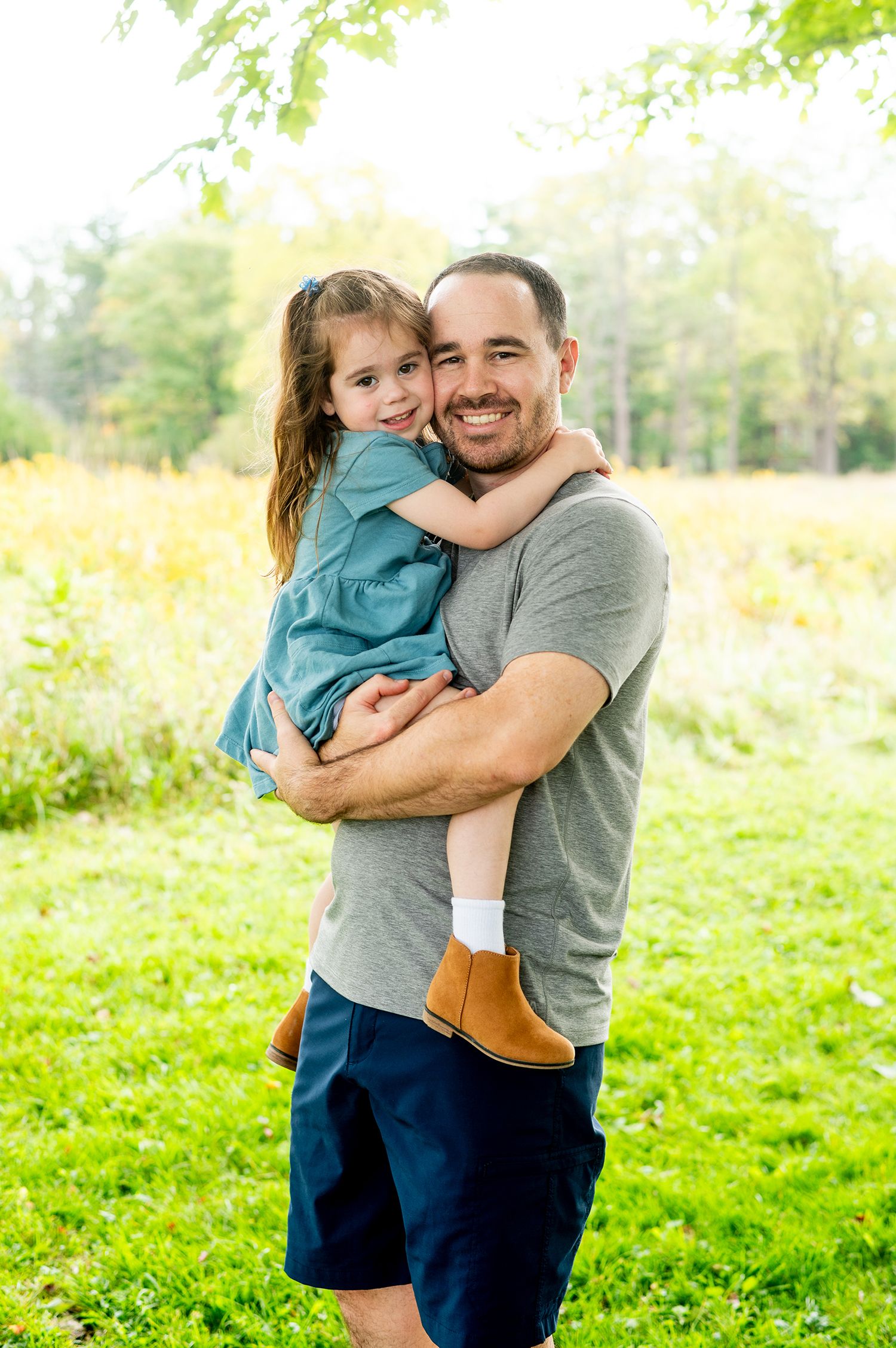 A smiling man holds a young girl in a blue dress outdoors in a lush green park setting.