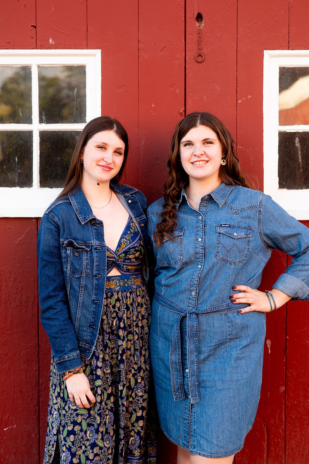 Two people wearing denim and floral clothing pose together against a rustic red barn wall with white windowpanes.
