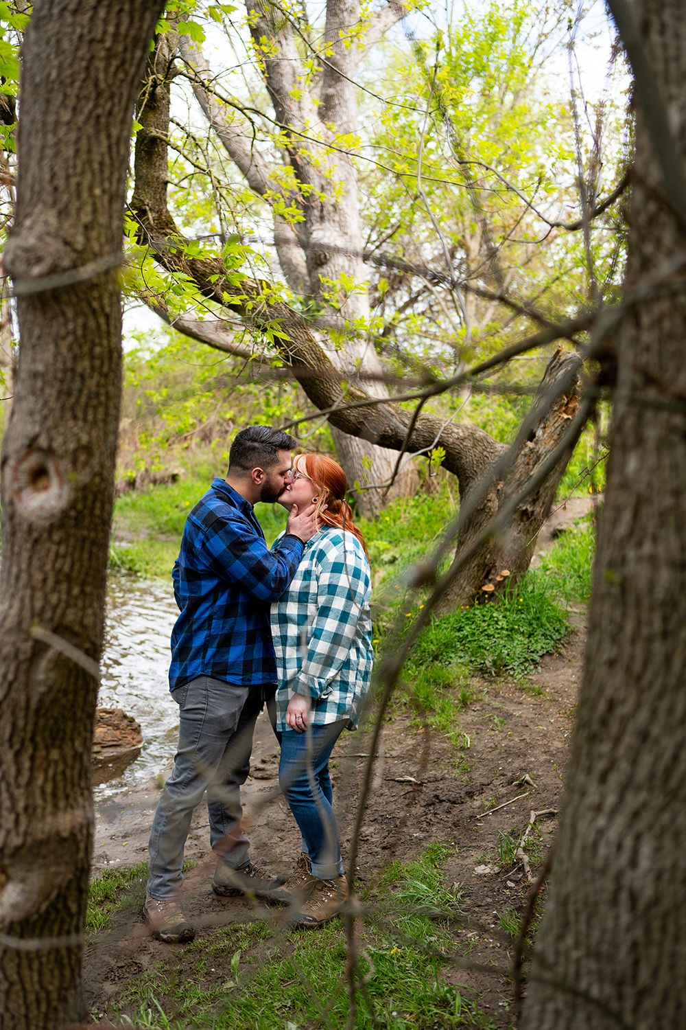A romantic couple in plaid shirts share kisses on a wooded trail by a stream during a fall engagement photo session.