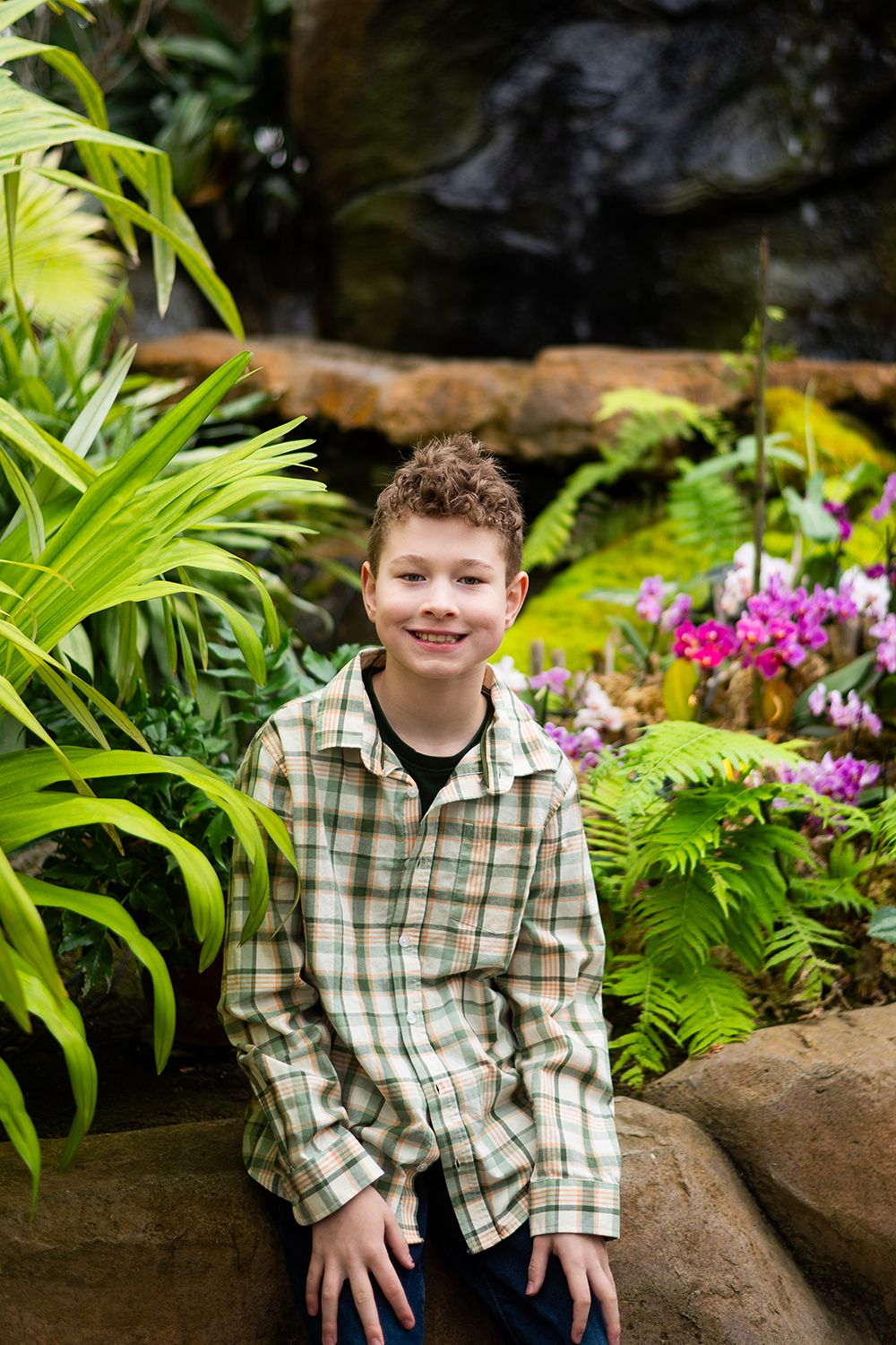 Child in plaid shirt sitting in a lush tropical garden setting with vibrant green plants and pink flowers.