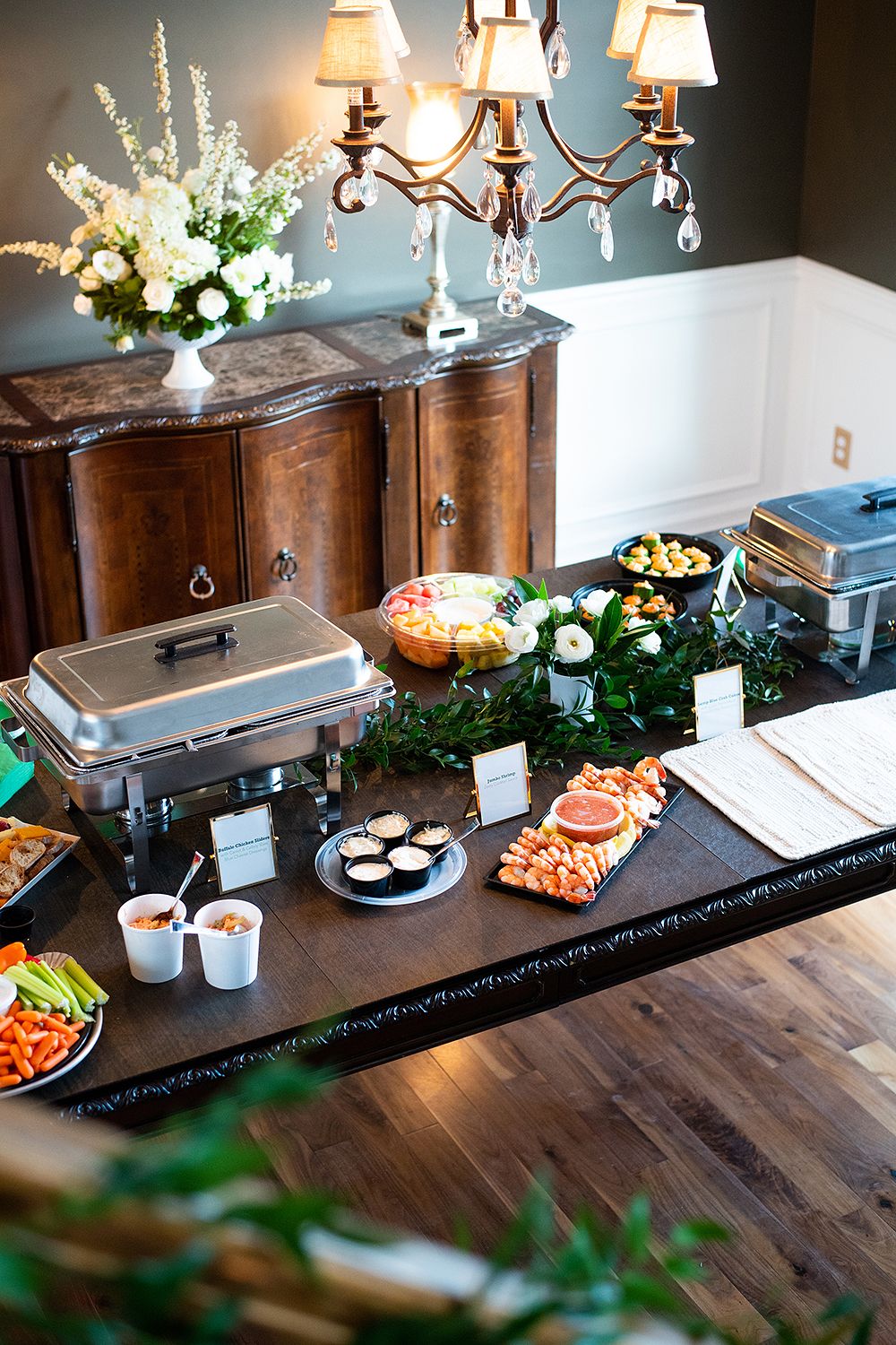 Elegant buffet table with chafing dishes and appetizer platters arranged on a dark wooden surface under warm chandelier lighting.