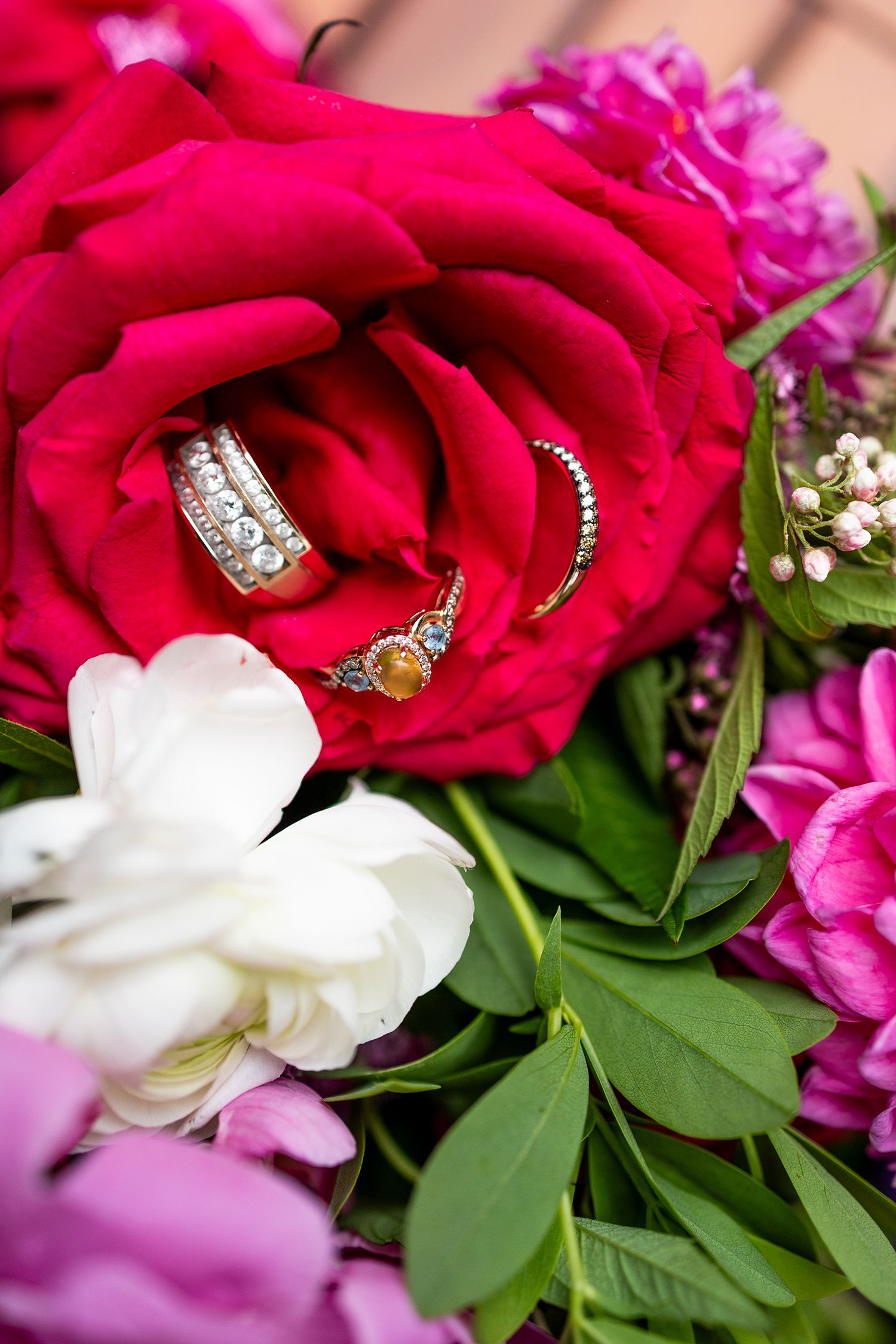 Elegant wedding rings nestled in a vibrant red rose surrounded by pink and white peonies in a bridal bouquet.