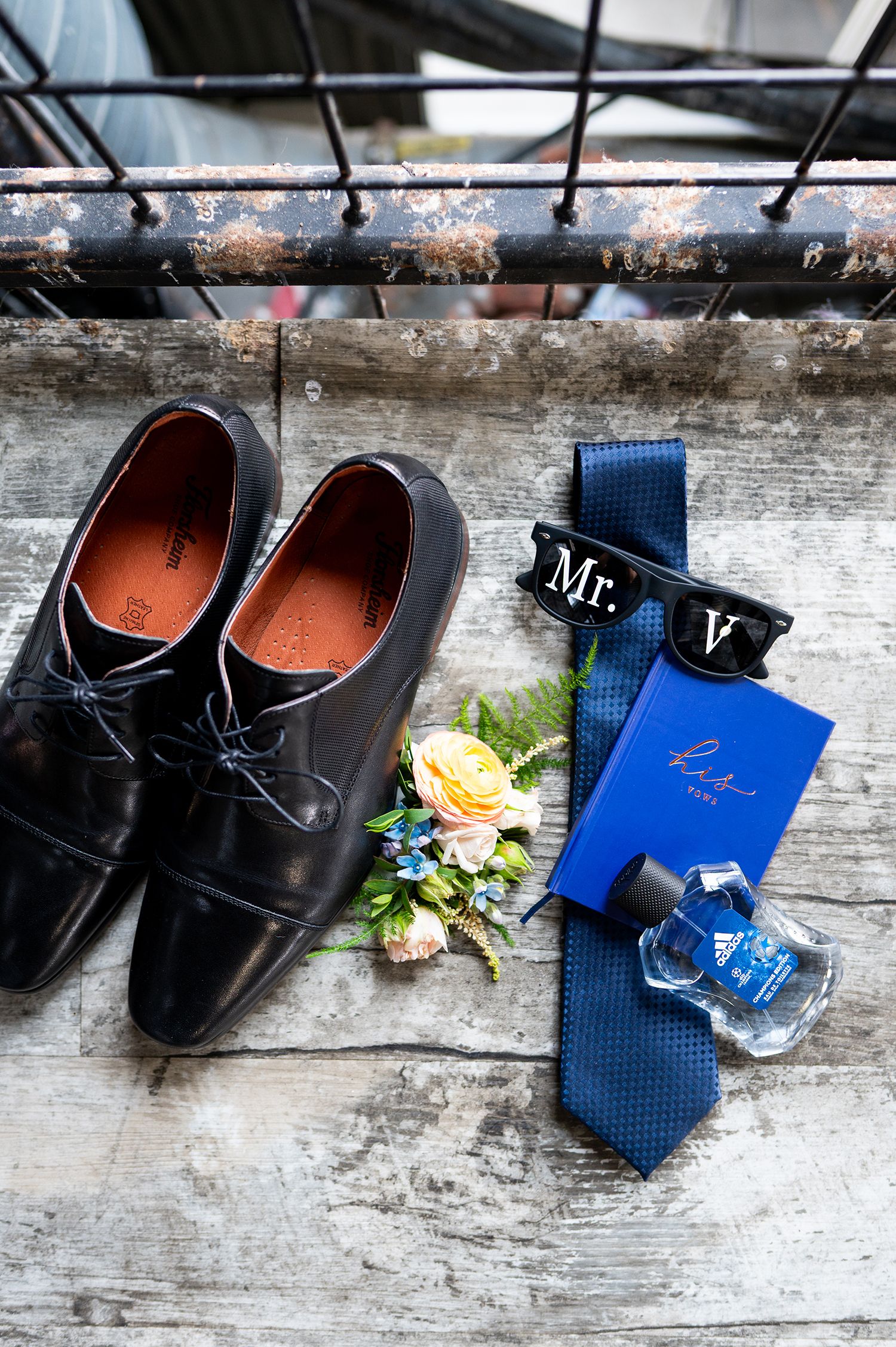 Black leather dress shoes with blue tie and scattered rose petals on a rustic wooden surface.