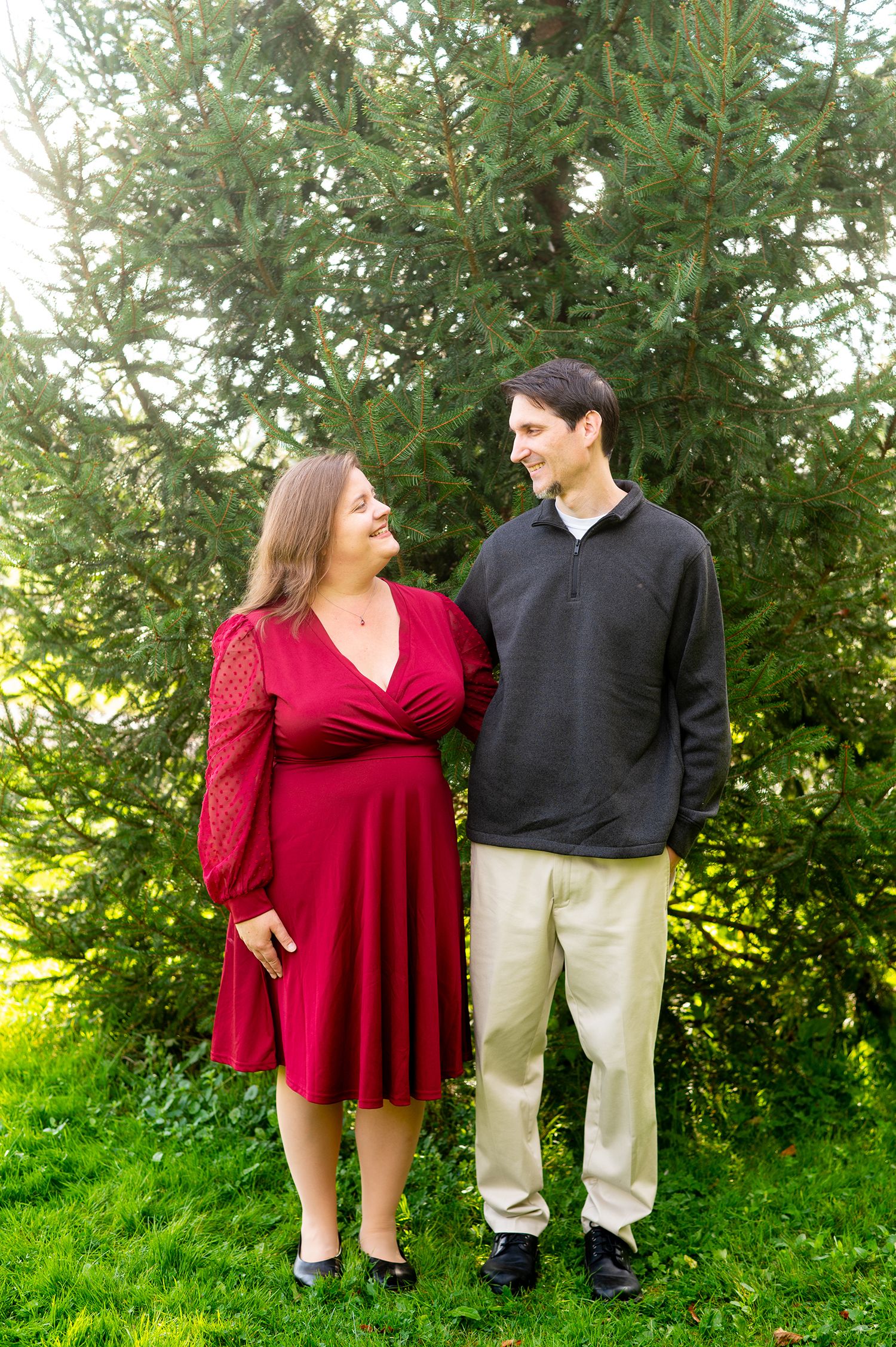 Couple smiling at each other outdoors, woman in red dress and man in black sweater, standing before lush green trees.