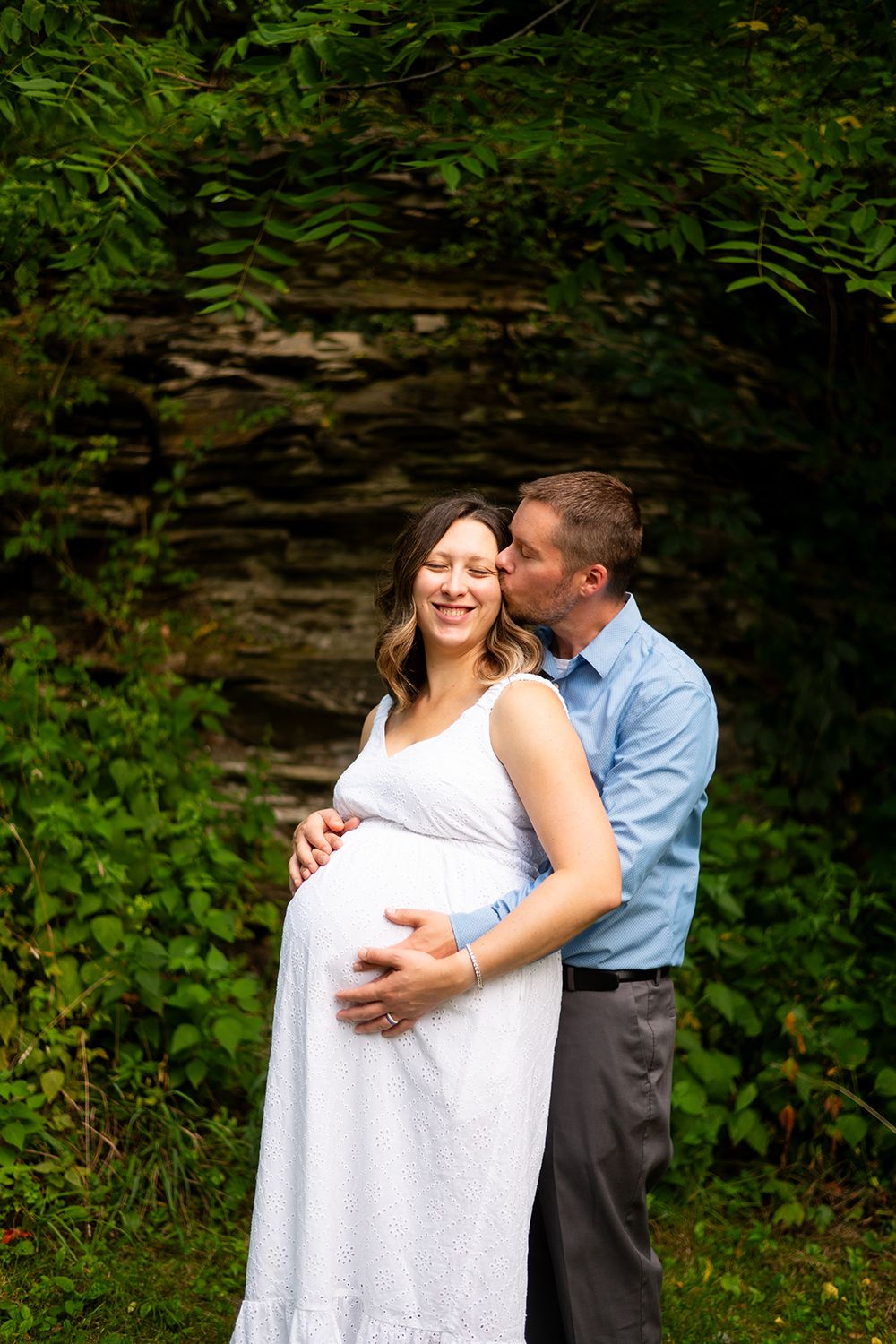 A couple shares an intimate moment during their maternity photo shoot in front of a stone wall and green foliage.