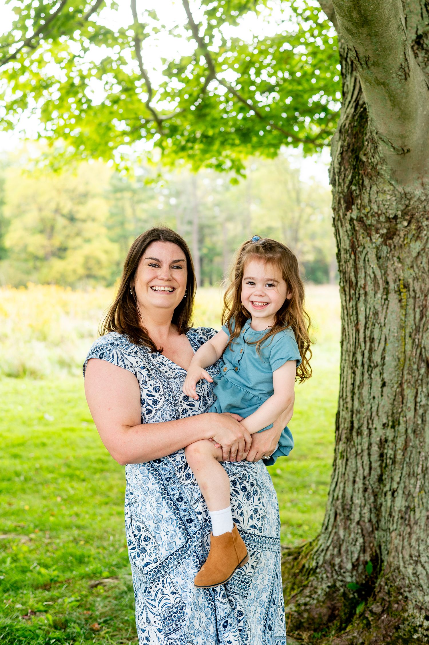A smiling woman in a blue floral dress holds a young girl in a teal outfit near a large tree in a lush green park.