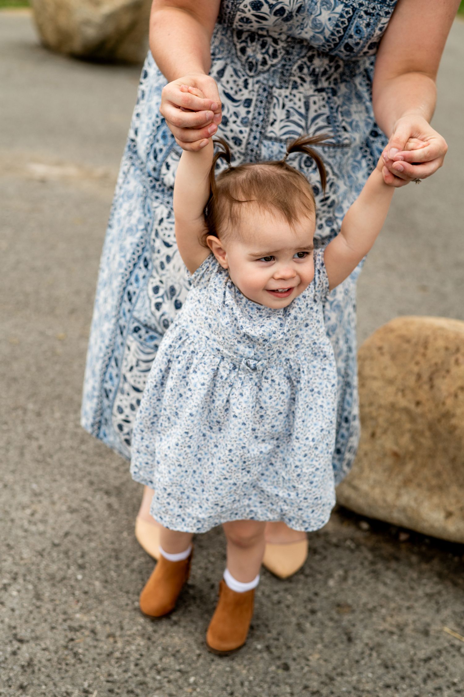 A toddler girl in a floral blue dress and brown boots takes first steps while an adult holds her hands.