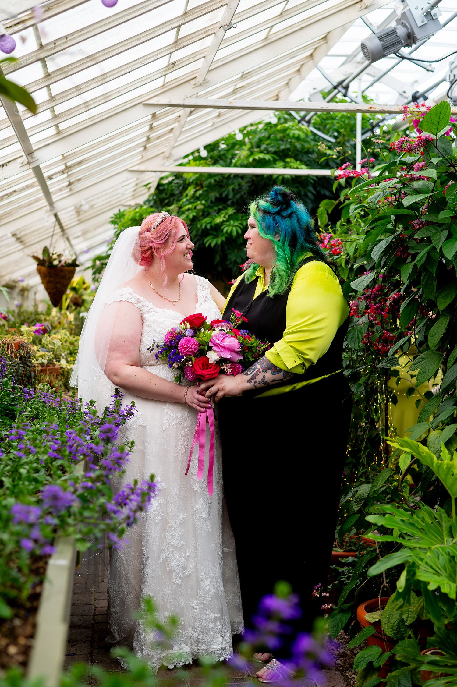 Two brides in a lush greenhouse, one in white lace with pink hair, the other in black and yellow with green hair, holding bouquet.
