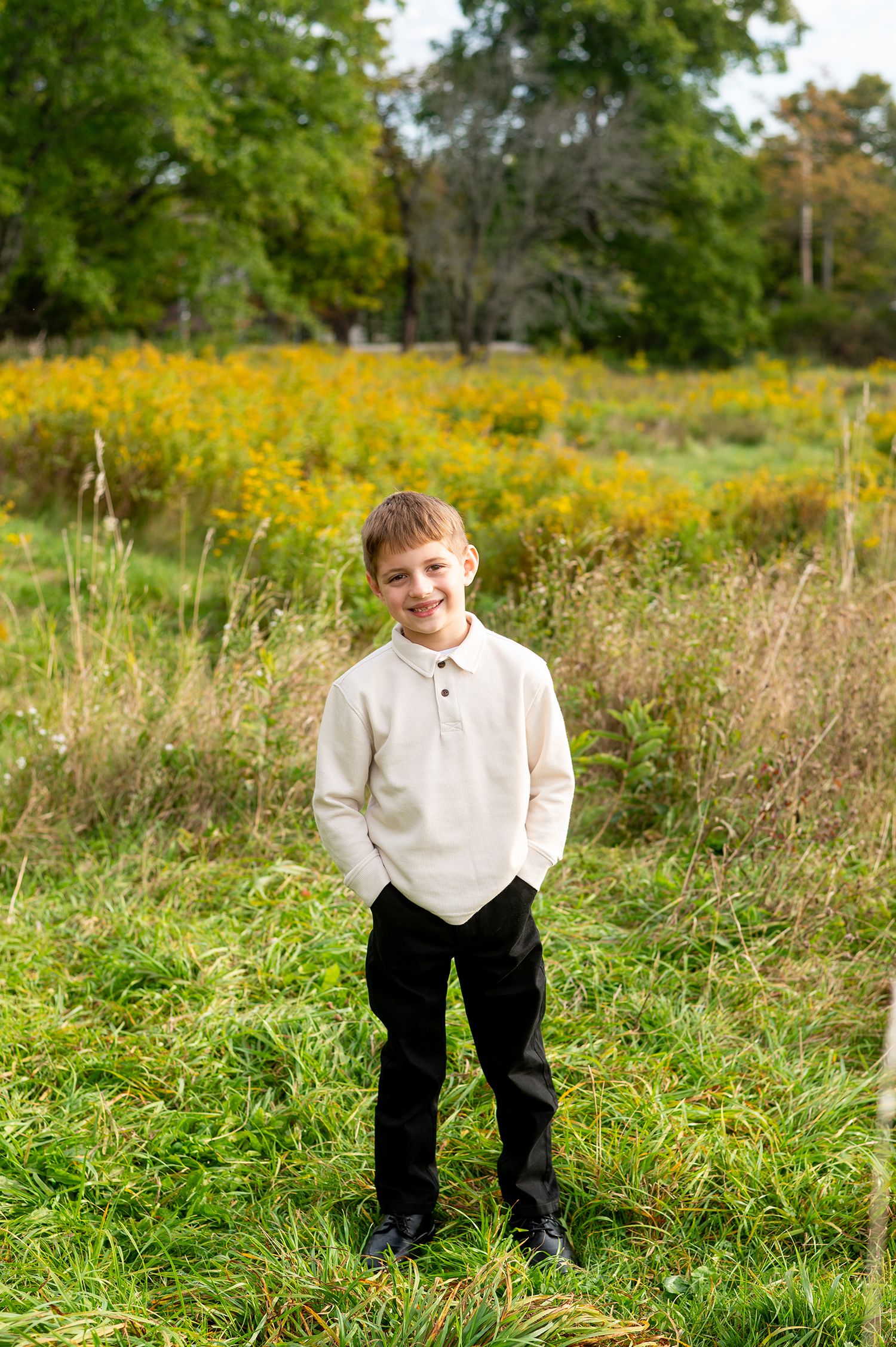 A smiling boy in a cream polo shirt and black pants stands in a lush green field with yellow wildflowers.