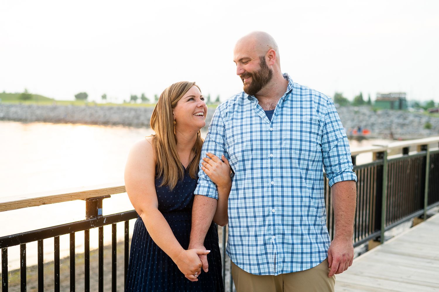 Happy couple holds hands and smiles on a waterfront bridge during a golden hour engagement photo session.