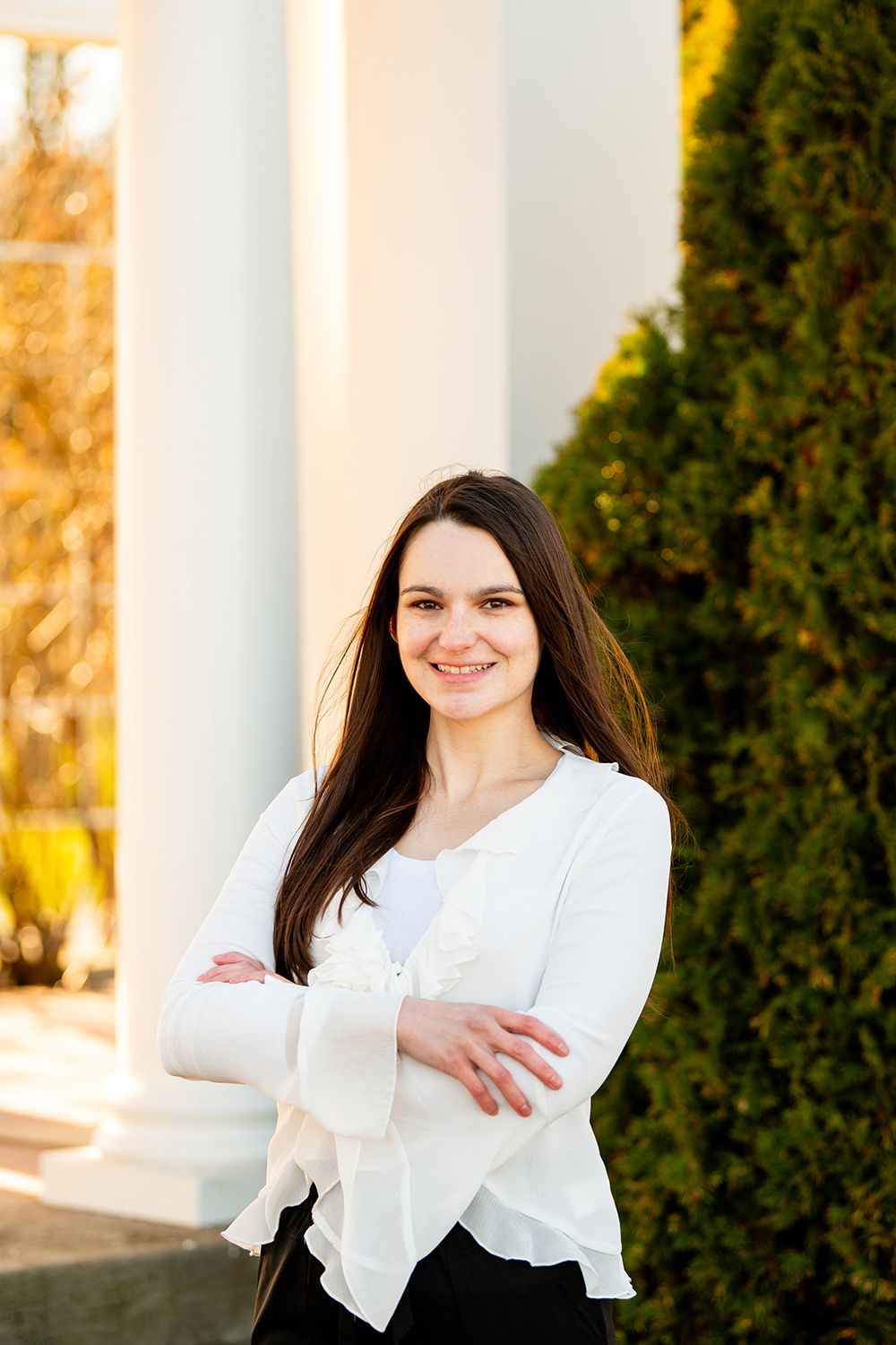 Portrait photo showing a person in white ruffled blouse smiling while standing against white columns with greenery.