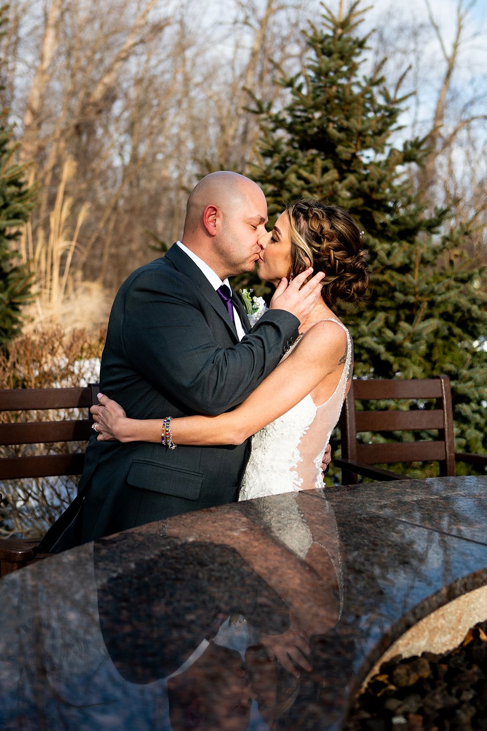 A romantic couple shares a tender kiss while seated on a bench with an evergreen tree in the background on a sunny day.
