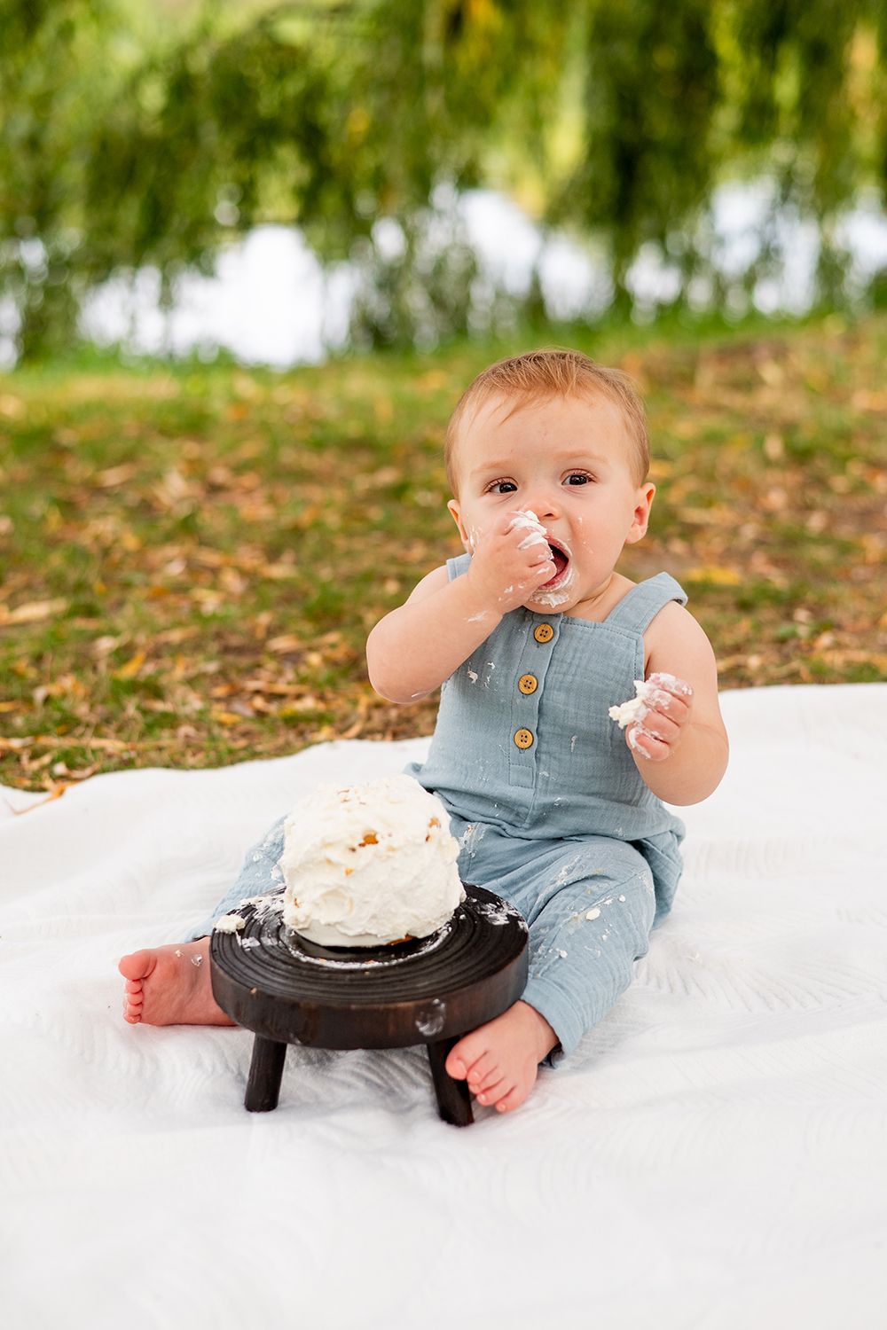 Baby in denim overalls sitting on white blanket having first birthday cake smash photo session in outdoor setting.