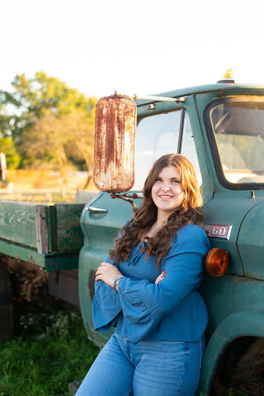 A person in a blue top poses against a vintage green farm truck in golden evening light.