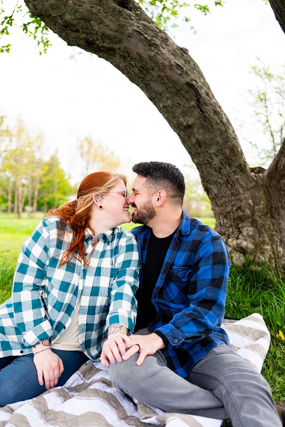 A couple in matching plaid shirts share an intimate moment while sitting under a curved tree trunk on a spring day.