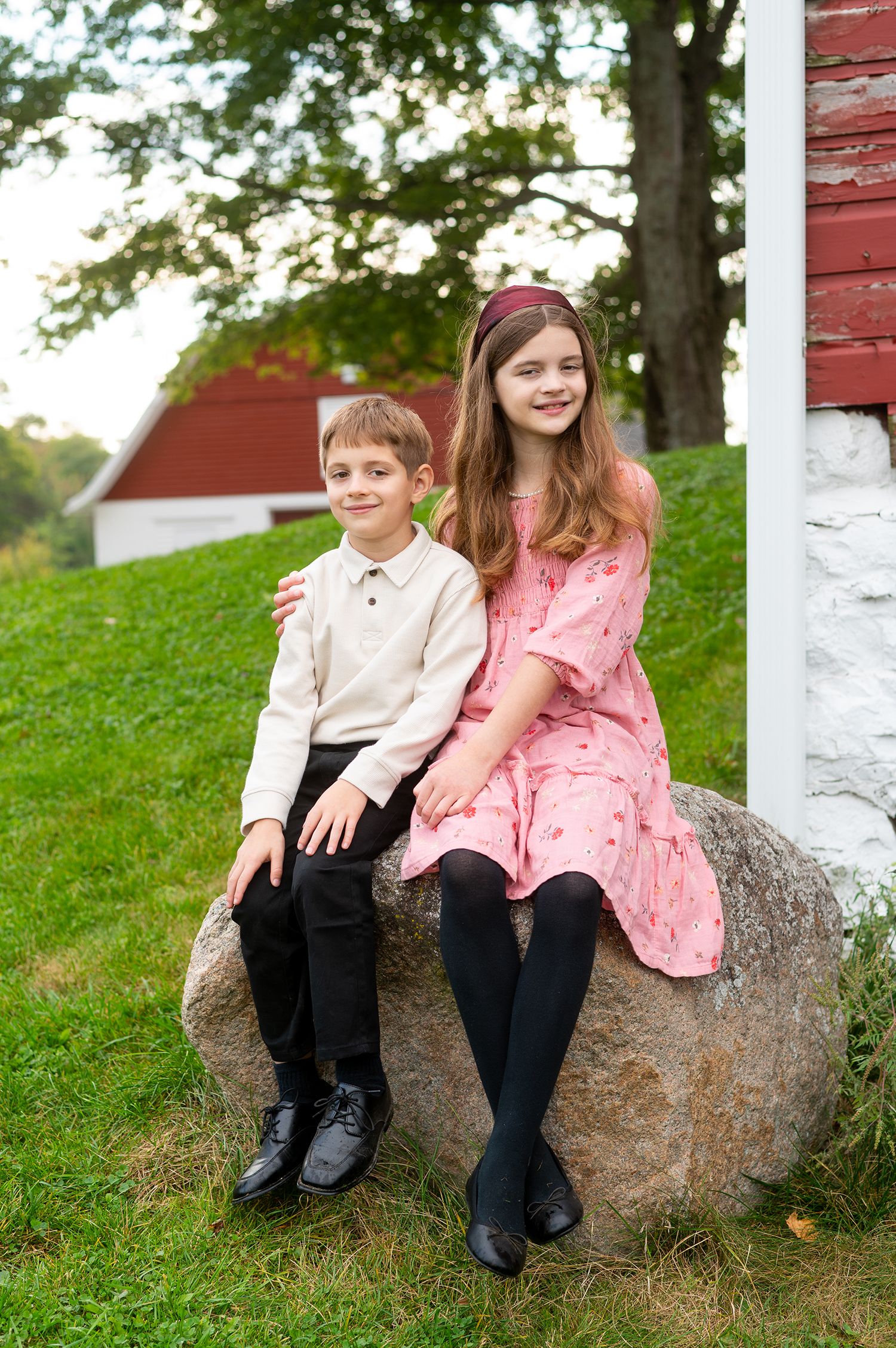 A smiling boy and girl in dressy clothes pose together on a rock by a red barn.