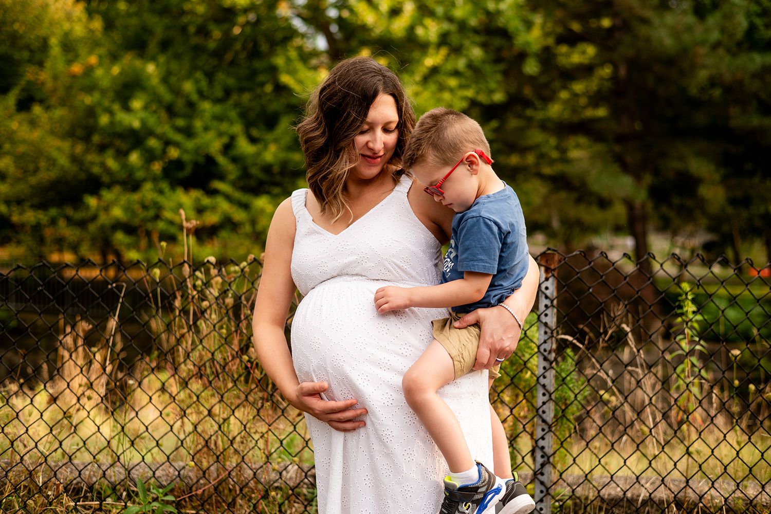 An expectant person in a white dress holds a young child outdoors in a natural setting during golden hour.