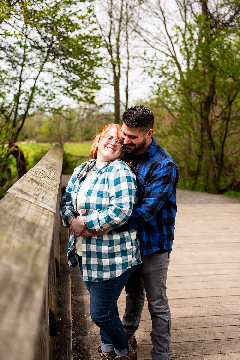 Romantic couple in matching blue plaid shirts embrace on a wooden boardwalk surrounded by spring foliage.
