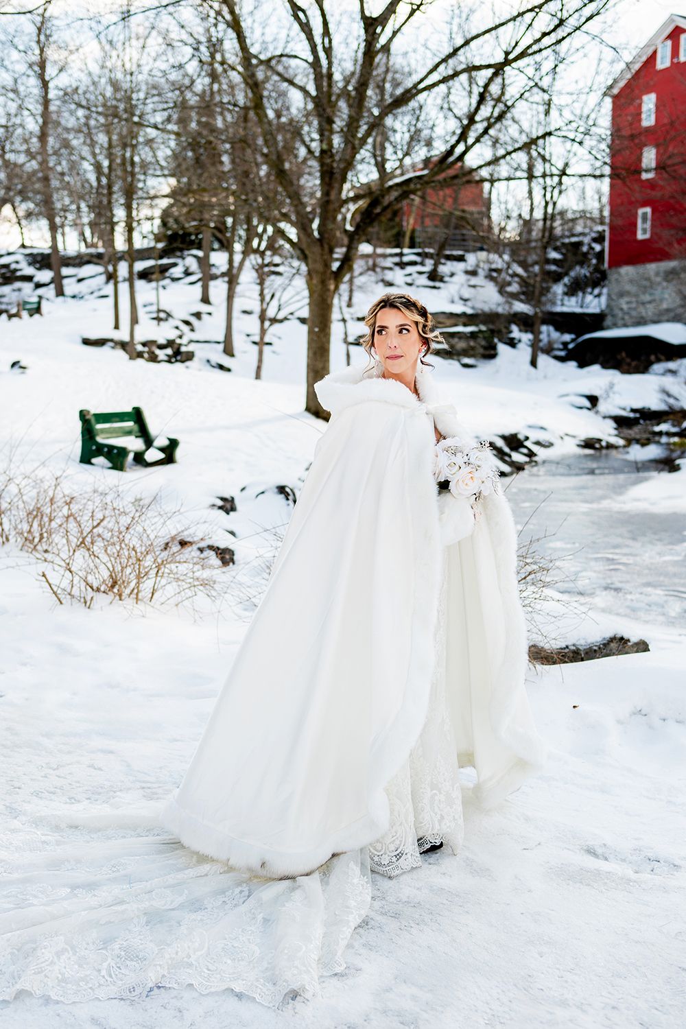 A white winter wedding gown with a cape stands out against snowy scenery with a red barn in the background.