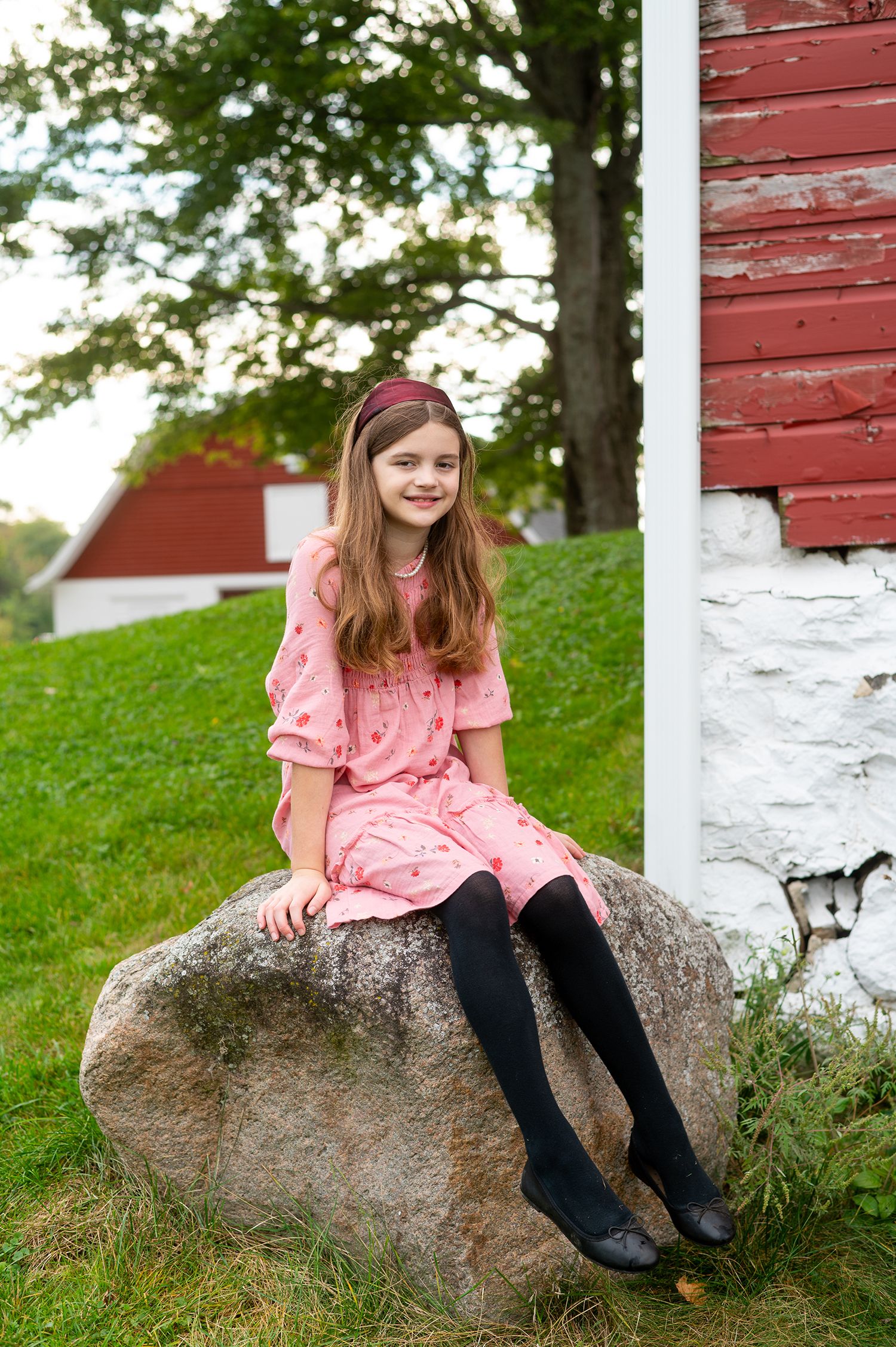 Young girl in pink dress and black tights sitting on a rock near a red barn, smiling at camera.