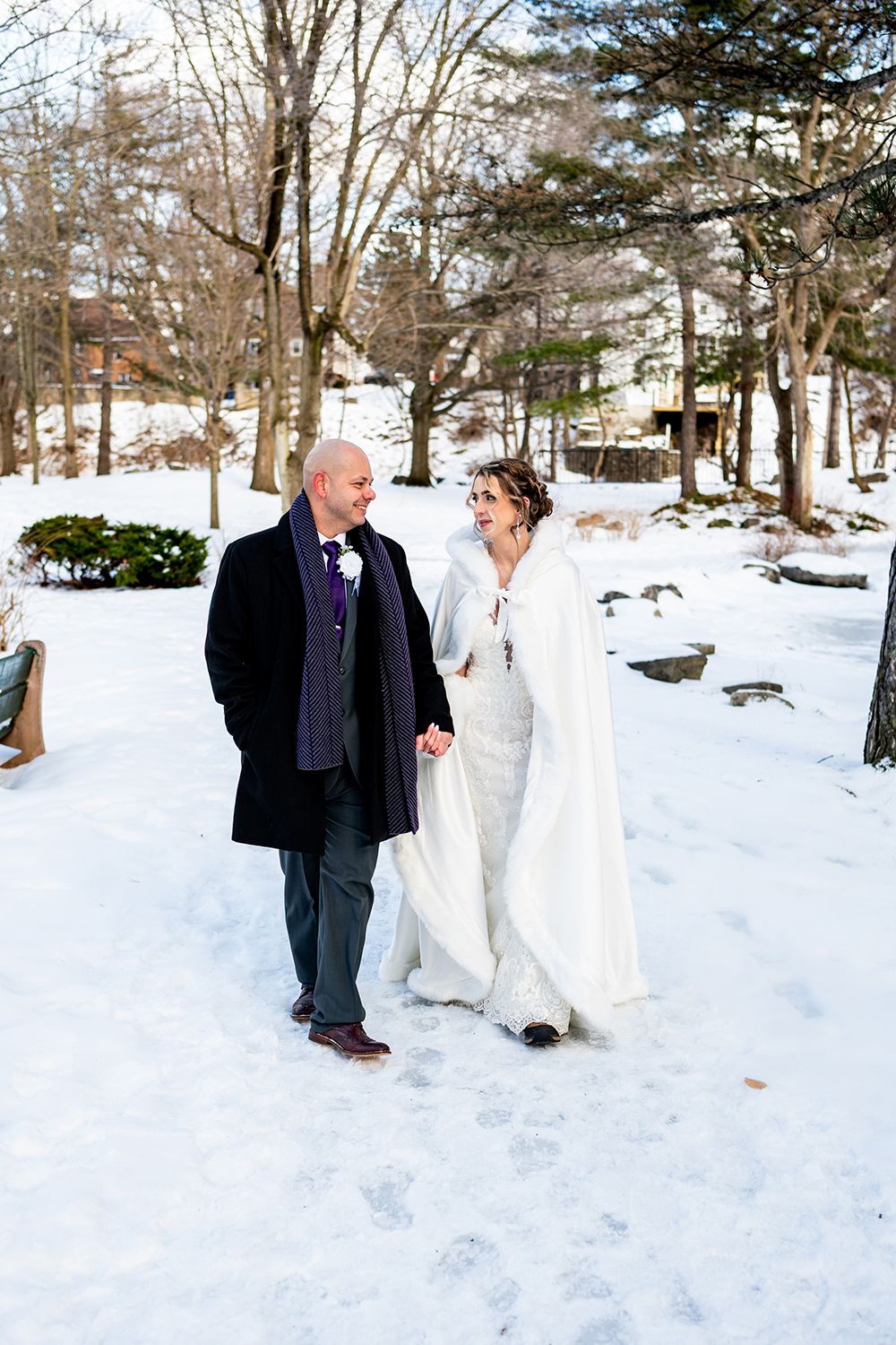 A couple walks together through a snowy winter landscape wearing formal attire for their wedding day.