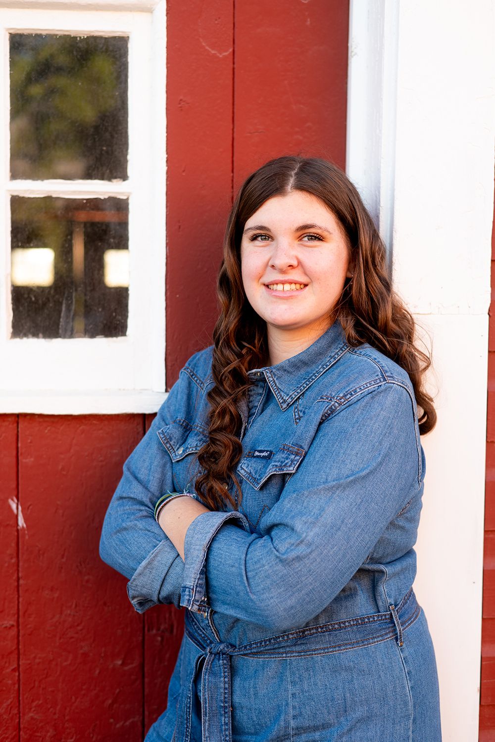 Someone wearing a denim jacket poses with arms crossed against a red and white barn wall with a window.