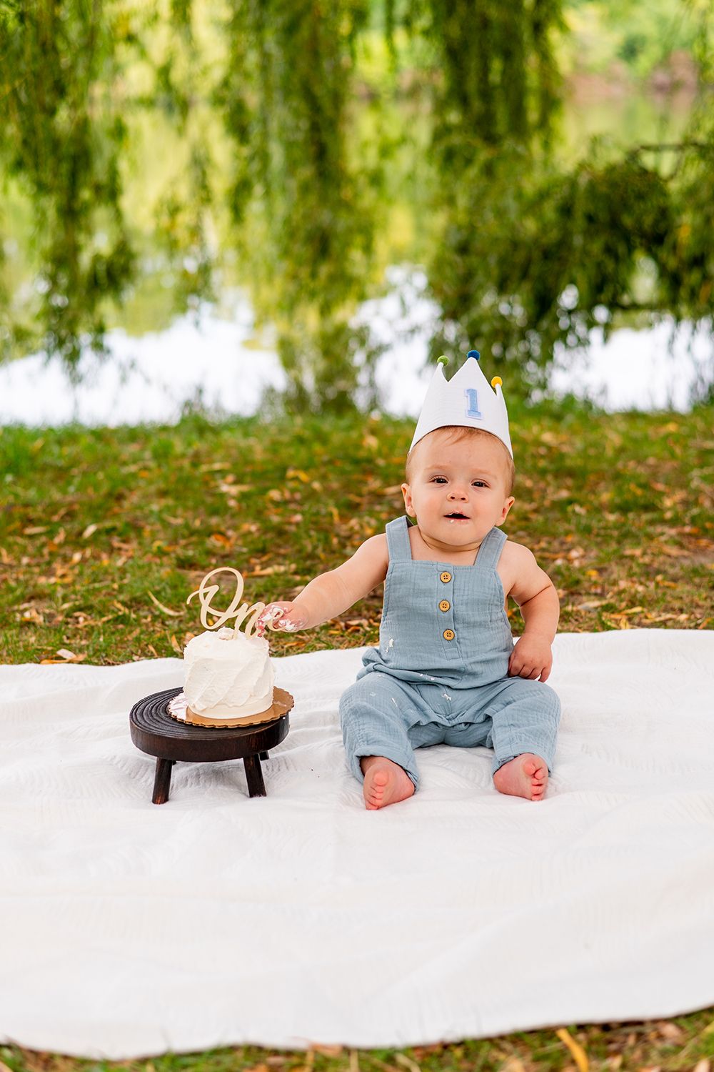 Baby wearing blue romper sits on white blanket by lake with small cake on wooden stool for first birthday celebration.