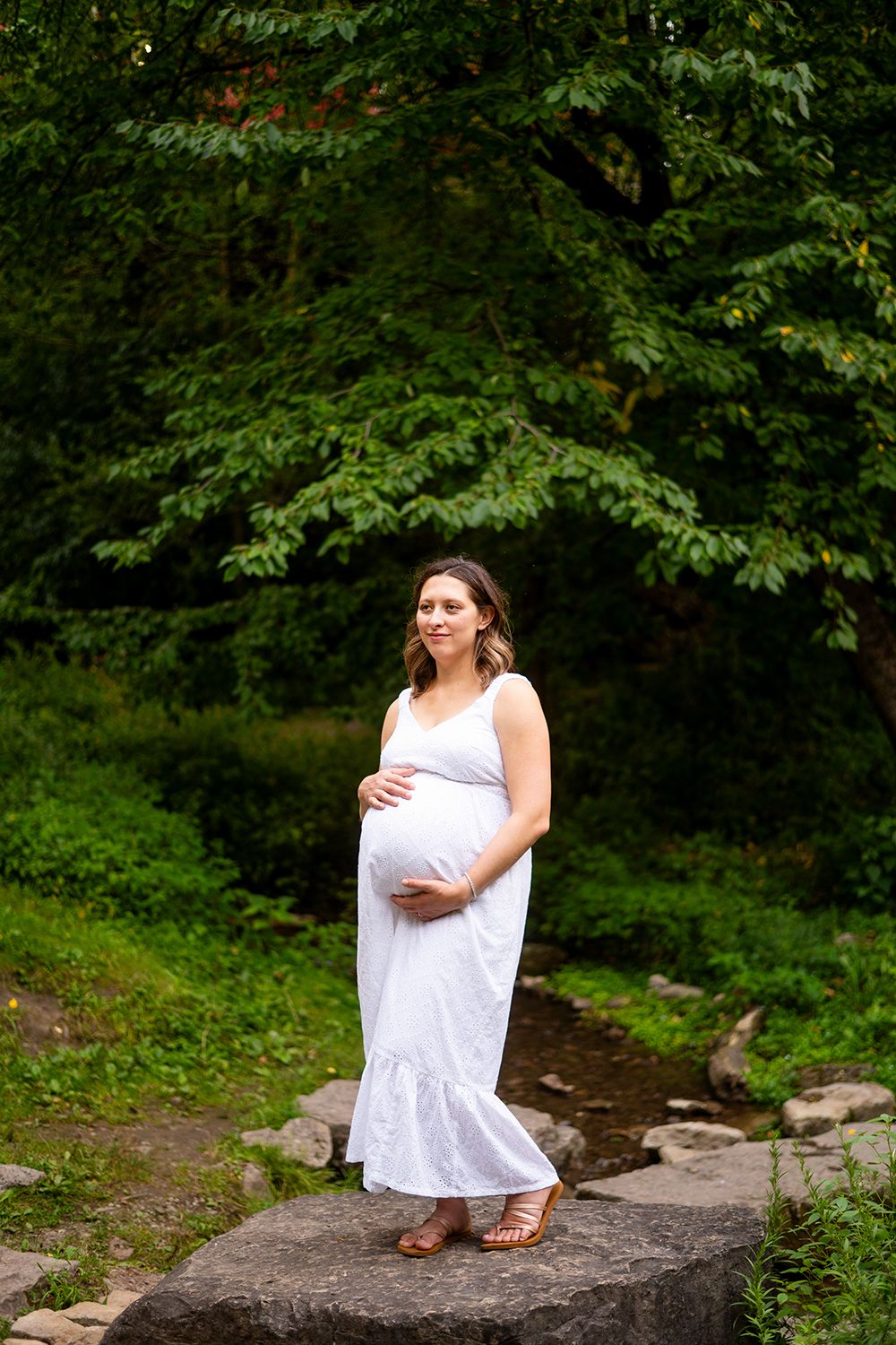 A pregnant woman in a white summer dress stands on a rock surrounded by lush green forest scenery.