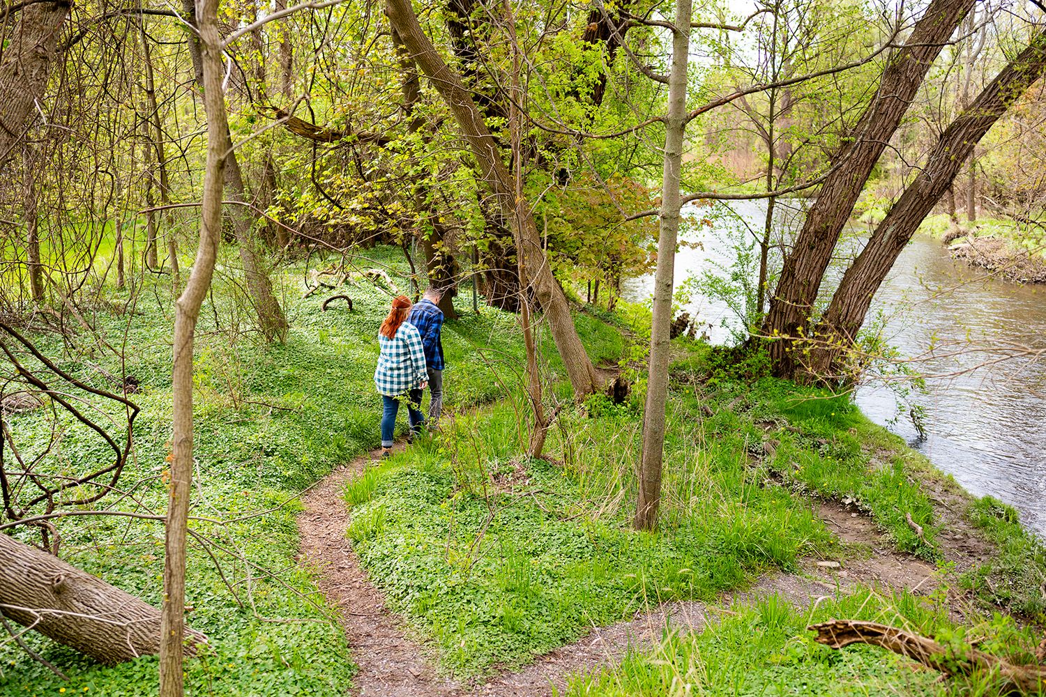 A hiker walks along a winding dirt trail through green woods beside a peaceful river on a spring day.