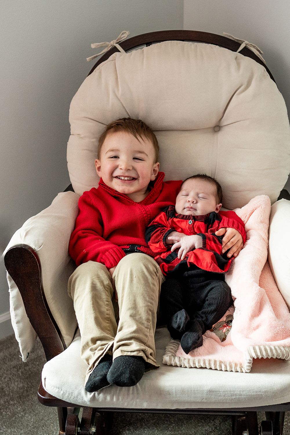 Two children in matching red outfits sit together on a cozy beige rocking chair.
