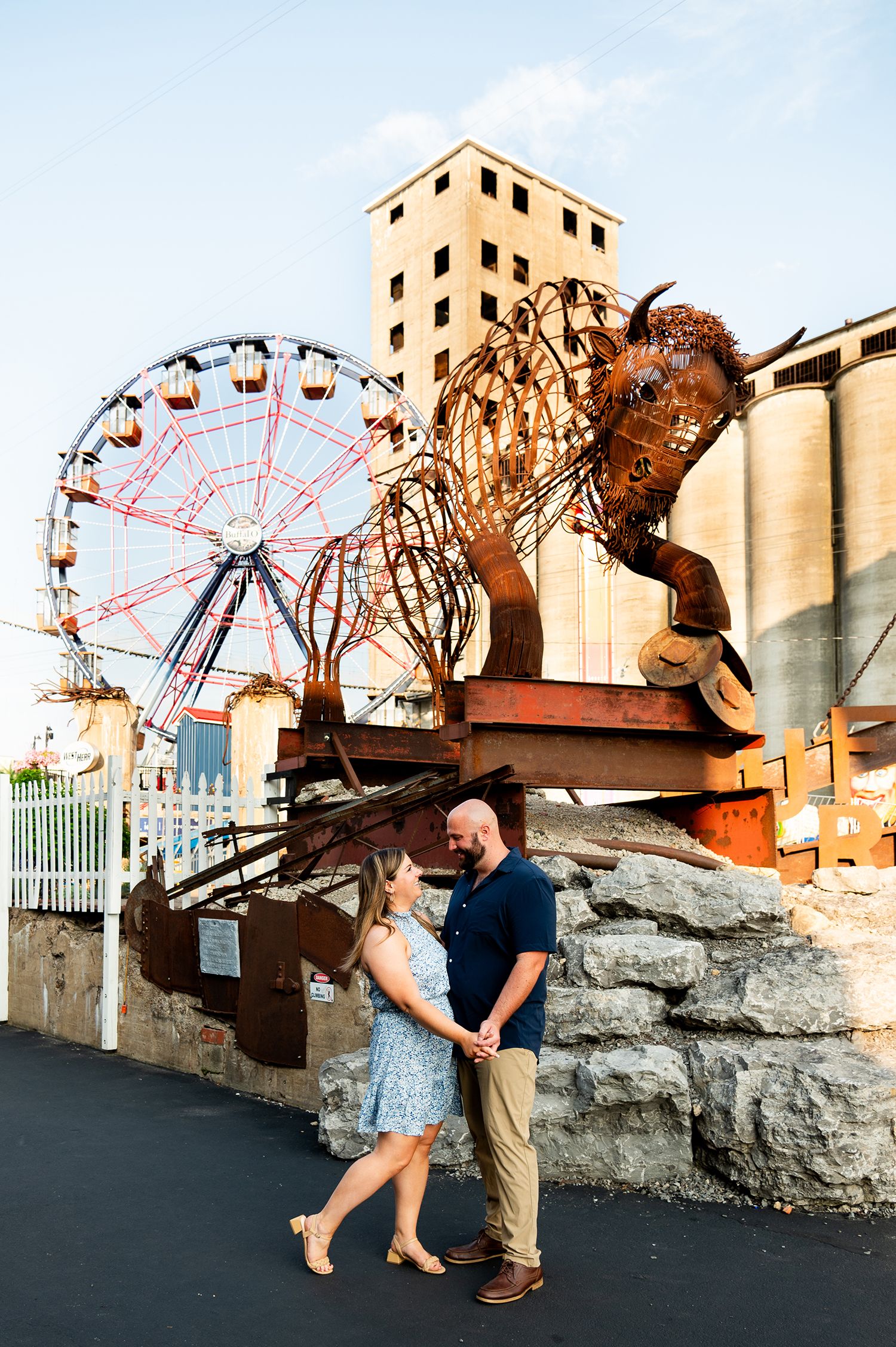 Couple holding hands near a large metal sculpture and Ferris wheel at an outdoor amusement area during golden hour.