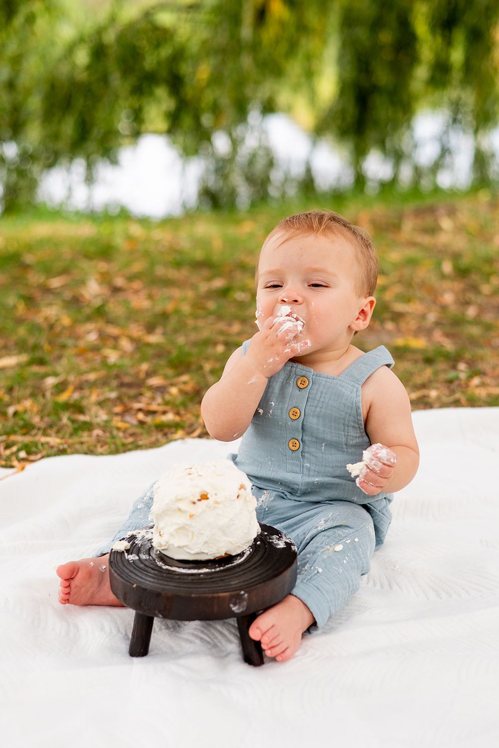 A small child in blue overalls sits on a white blanket enjoying a cake smash photoshoot outdoors.