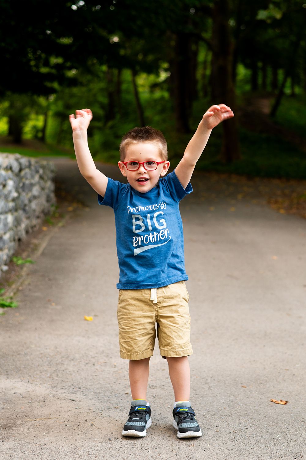 Young child in glasses wearing a 'Big Brother' shirt raises arms joyfully while standing on a path.