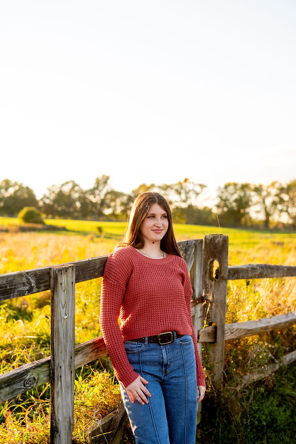 Person in a red sweater and jeans poses casually by a rustic wooden fence at sunset in a golden field.