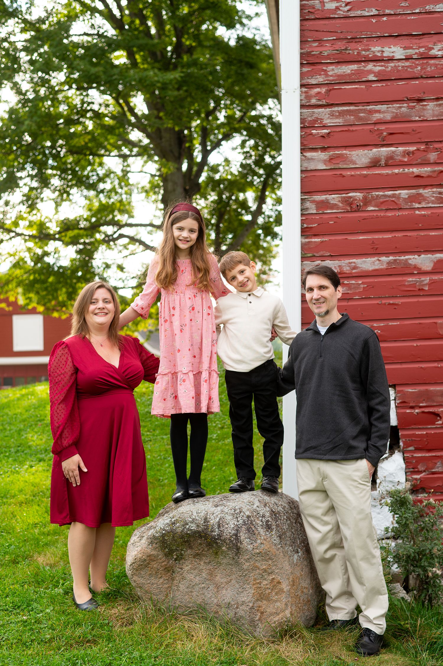 Family of four posing outdoors near a red barn, two children standing on a rock between smiling parents.
