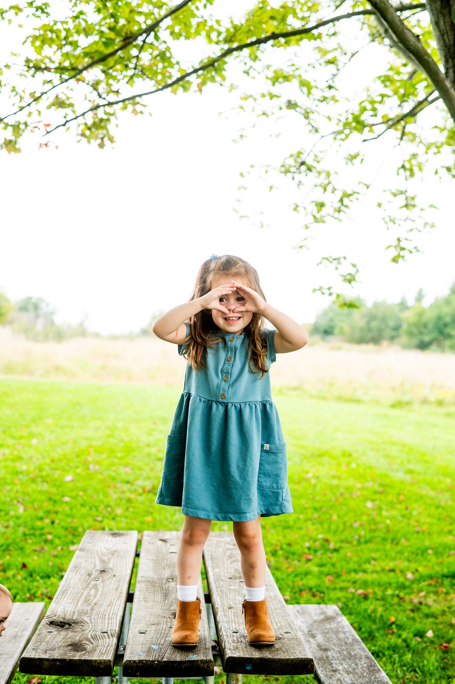A young girl in a teal dress and brown boots stands on a picnic table, making binocular shapes with her hands over her eyes.