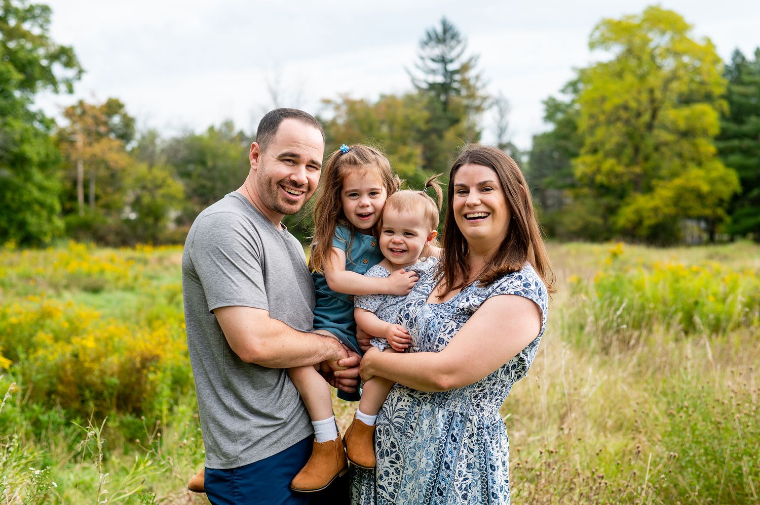 Happy family of four posing outdoors in a meadow, parents holding two young daughters, smiling in a scenic field.