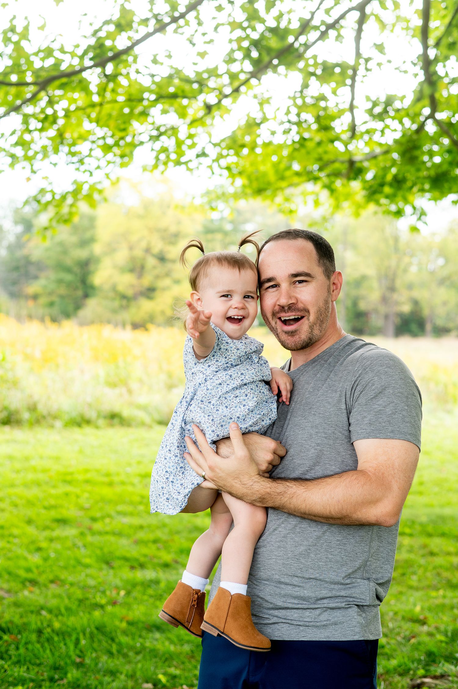 A joyful father holds his laughing baby girl in a sunny green park, both smiling brightly outdoors.