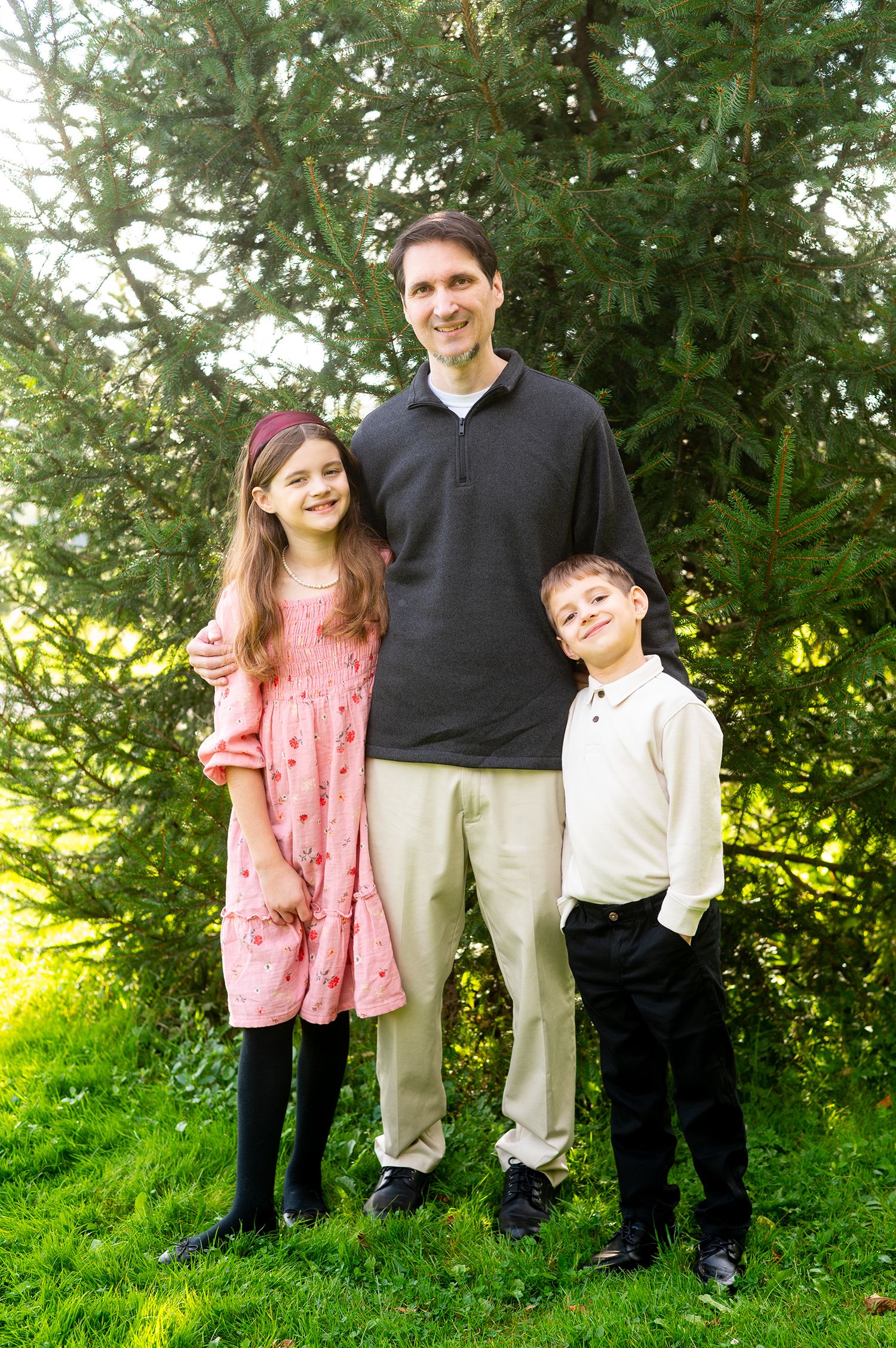 A smiling father poses outdoors with his daughter in a pink dress and son in white shirt amid lush green trees.