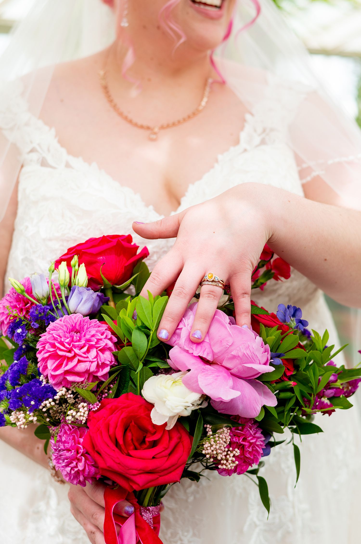 Bride in white lace dress displaying wedding ring over colorful bouquet of red roses, pink peonies, and purple flowers.