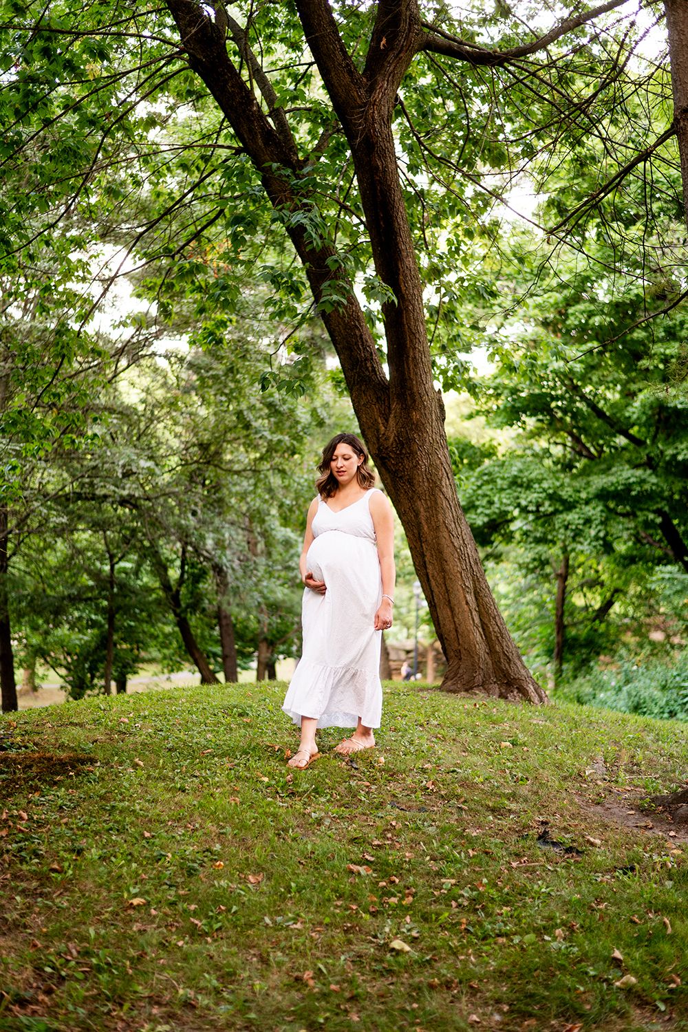 A figure in a flowing white dress stands beneath a large tree in a lush green park setting.