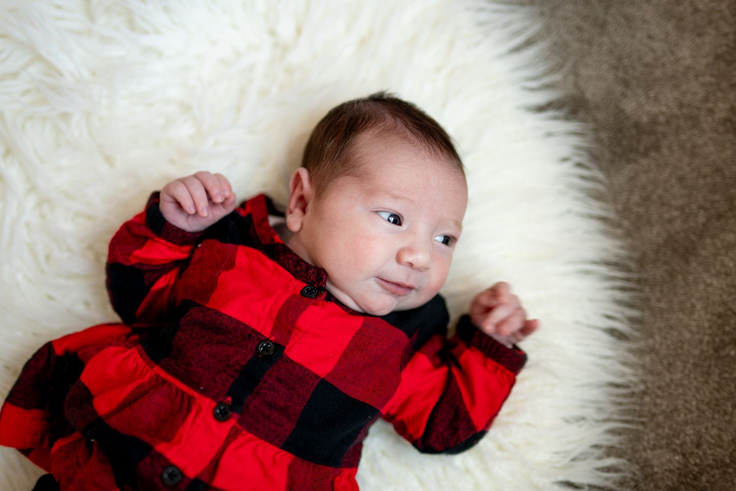 Baby wearing red and black buffalo plaid outfit lies on white furry surface.