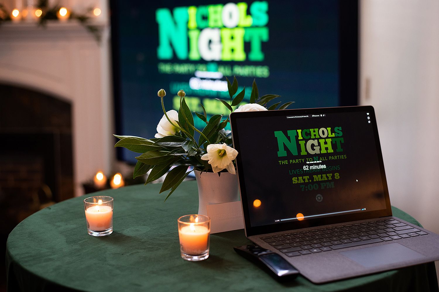A laptop displays 'Trivia Night' in green neon text on a dark table with white flowers and glowing candles.