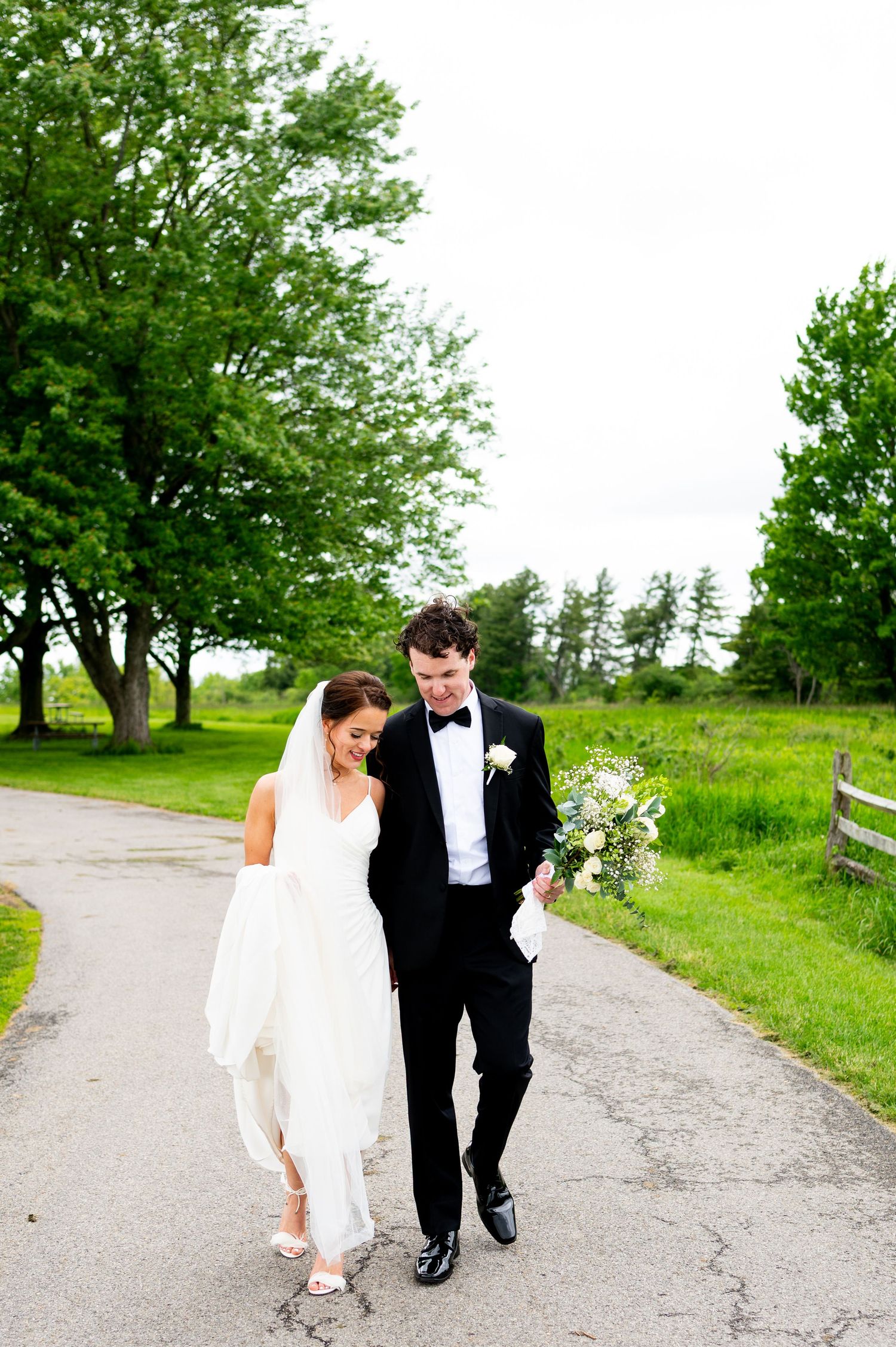 A couple in formal wedding attire walks along a scenic country path surrounded by lush green trees and grass.