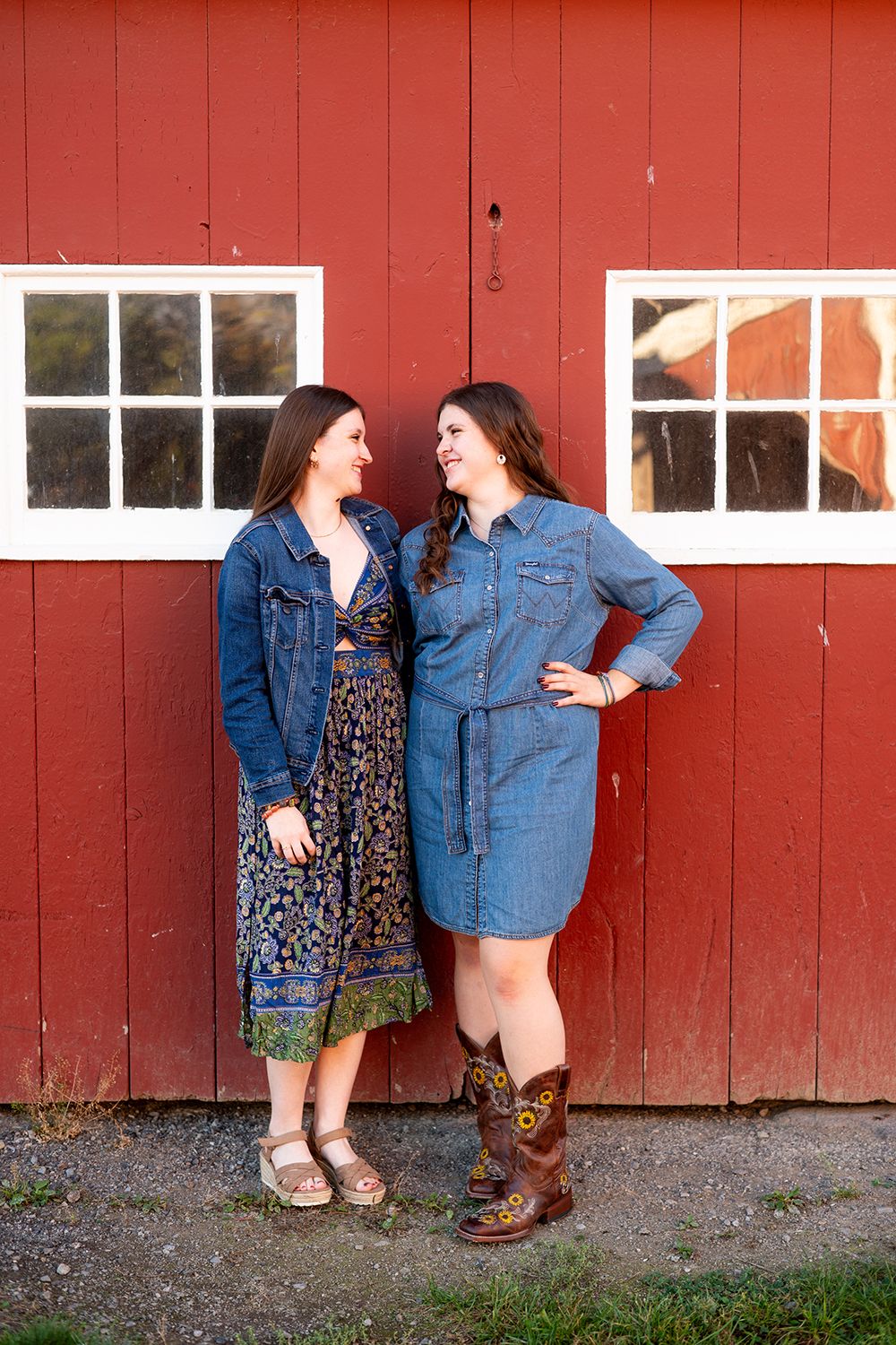 Two friends laugh together while posing against a bright red barn wall, wearing denim and cowboy boots.