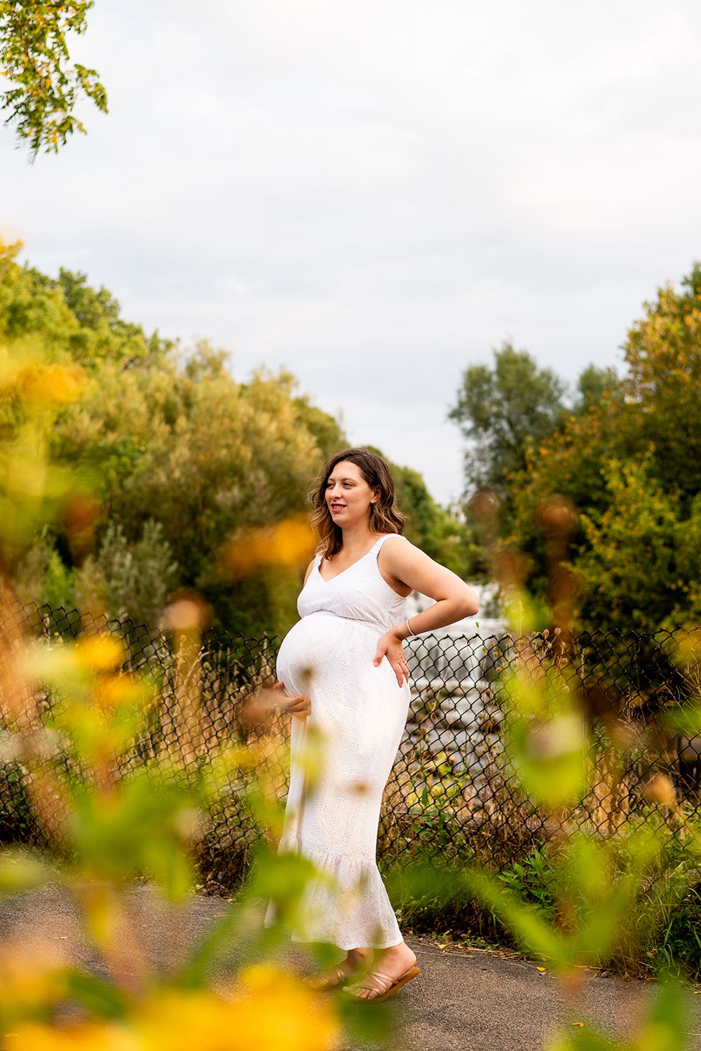 Figure in white dress poses gracefully against rustic fence with autumn foliage creating dreamy bokeh effect.